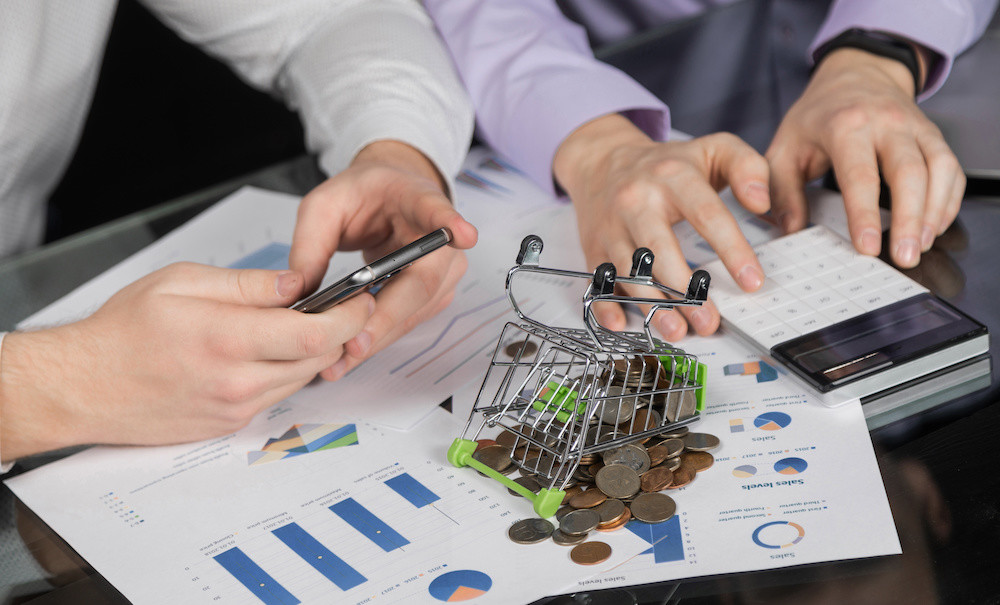 Two men is using his hands on calculator for calculate products online price which have coins in shopping trolley beside laptop on office table. Online shopping and e-commerce concept