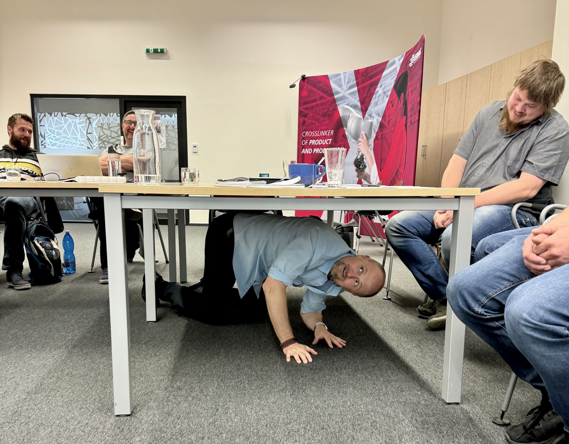 Lektor tréninku projektového řízení s nadsázkou předvádí cvičení pod stolem, zatímco účastníci se smějí. Atmosféra tréninku je uvolněná a přátelská.
