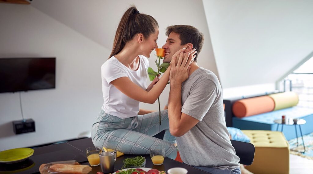 Mladý pár se směje u snídaně, žena drží růži a muž se usmívá, symbol každodenní radosti, blízkosti a lásky.