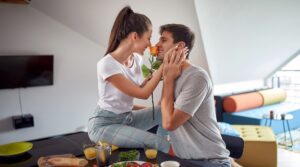 Mladý pár se směje u snídaně, žena drží růži a muž se usmívá, symbol každodenní radosti, blízkosti a lásky.