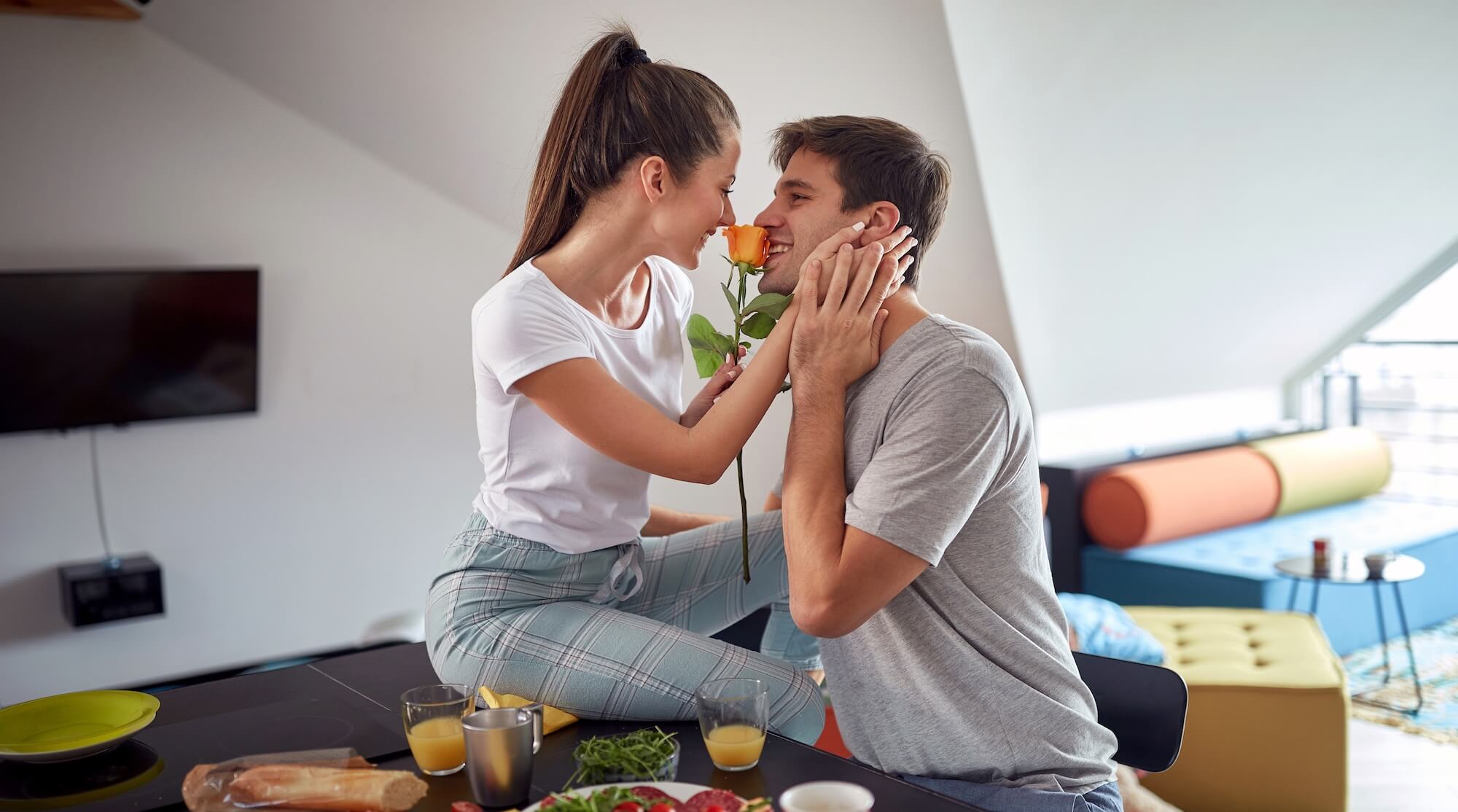 Mladý pár se směje u snídaně, žena drží růži a muž se usmívá, symbol každodenní radosti, blízkosti a lásky.